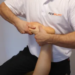 Masaż stopy wykonywany przez osobę w białej koszulce z logo 'FIZIOBALANS', widoczne czarne spodnie i fragment ściany.