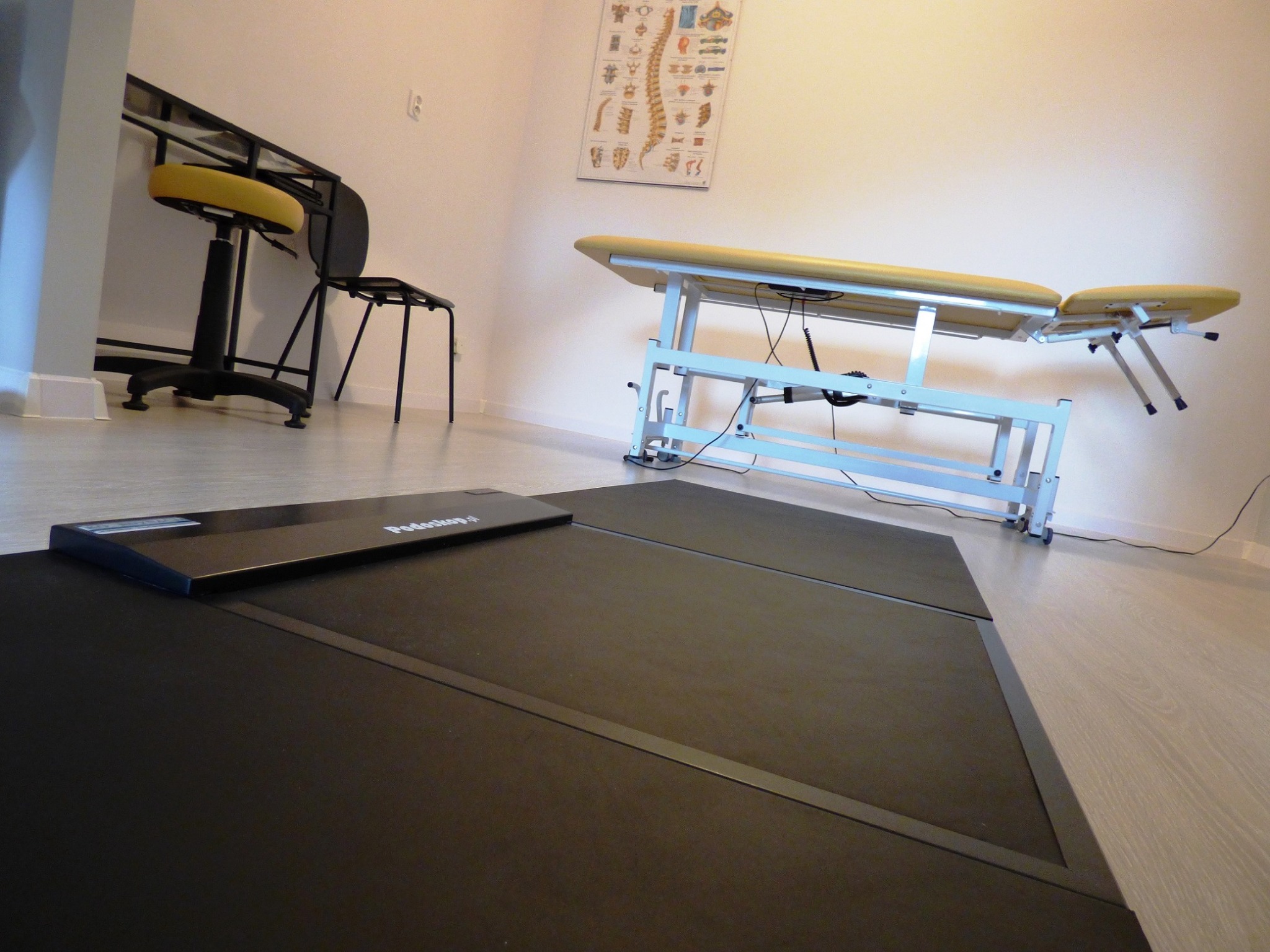 Gabinet: Stół do terapii z żółtym obiciem, plakat z kręgosłupem na ścianie, czarna platforma diagnostyczna Podostop i krzesło obrotowe w tle.