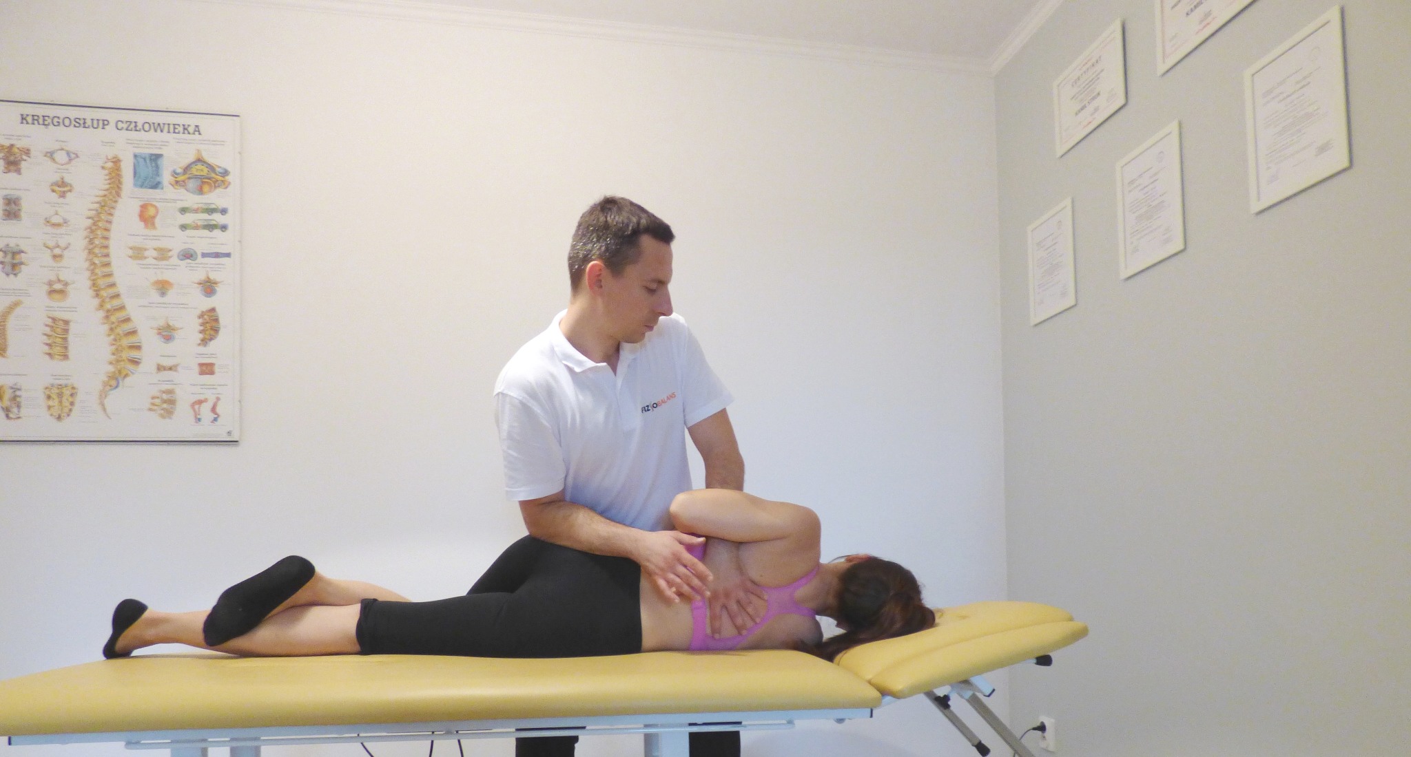 Pacjentka leżąca na boku na żółtym stole do masażu podczas terapii manualnej kręgosłupa przez terapeutę w białej koszulce z logo, na ścianie plakat z budową kręgosłupa i certyfikaty.