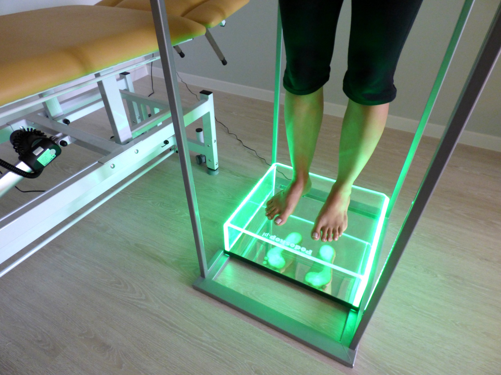 Pacjentka na podoskopie z zielonym podświetleniem, widoczne odciski stóp, w tle kozetka rehabilitacyjna.