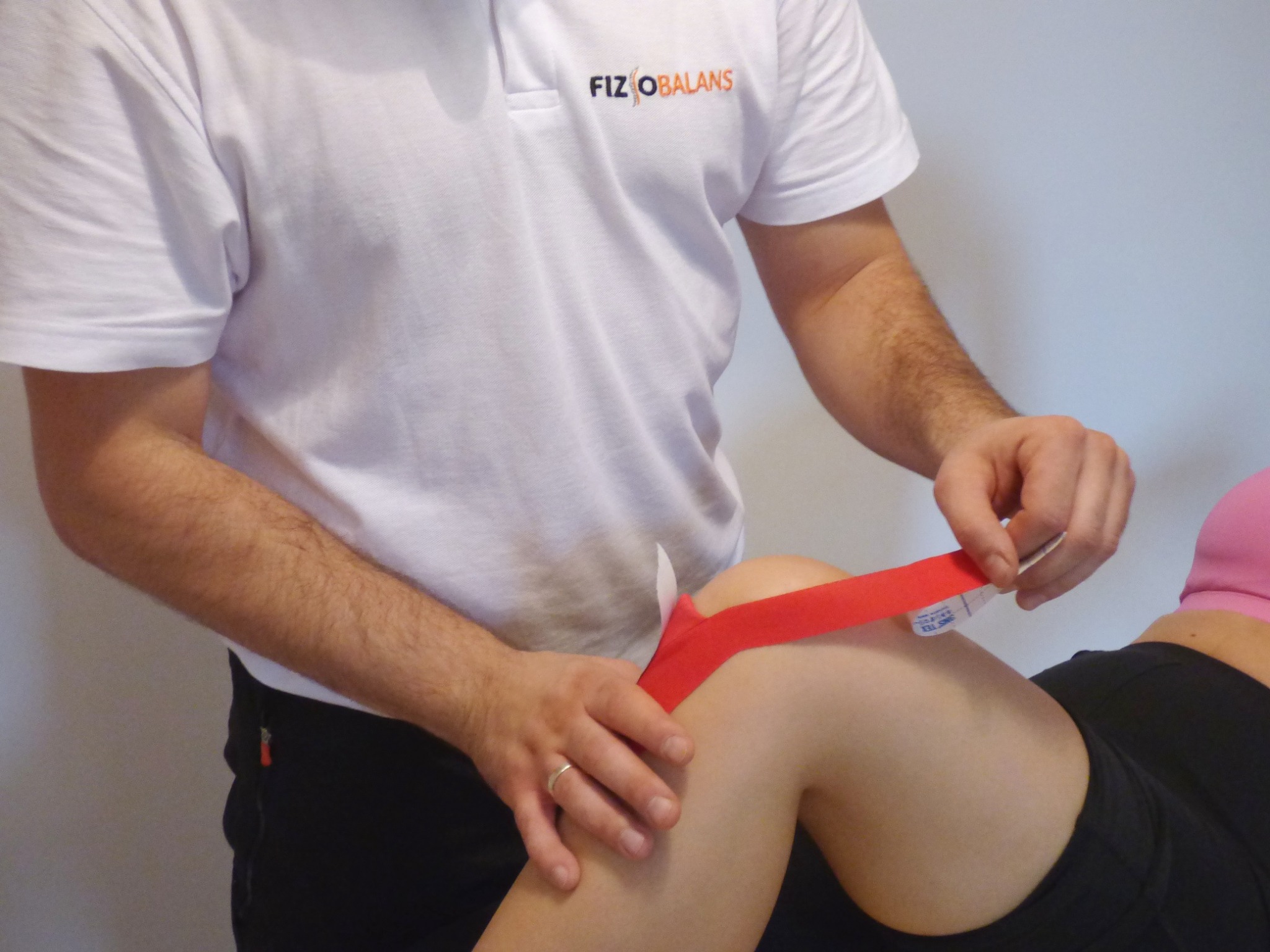 Naklejanie czerwonej taśmy kinesiotaping na kolano pacjentki przez terapeutę w białym polo z logo FIZIOBALANS.
