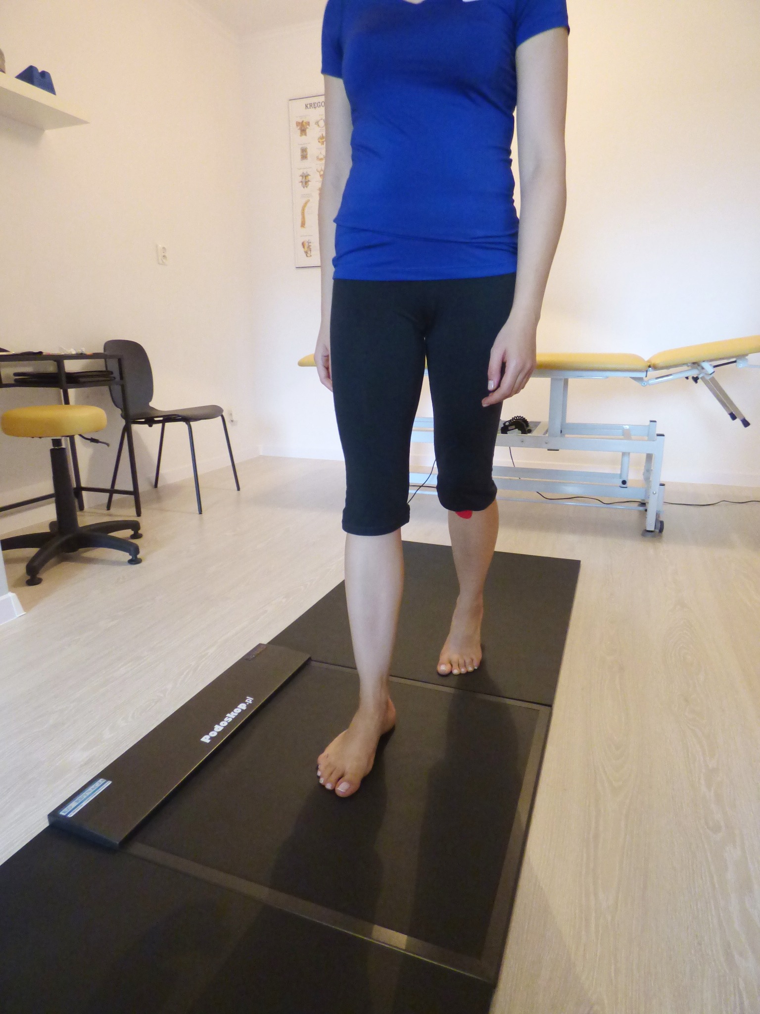 Pacjentka stąpa boso po platformie podoskopowej w gabinecie, widoczny plakat z układem kostnym na ścianie i kozetka.