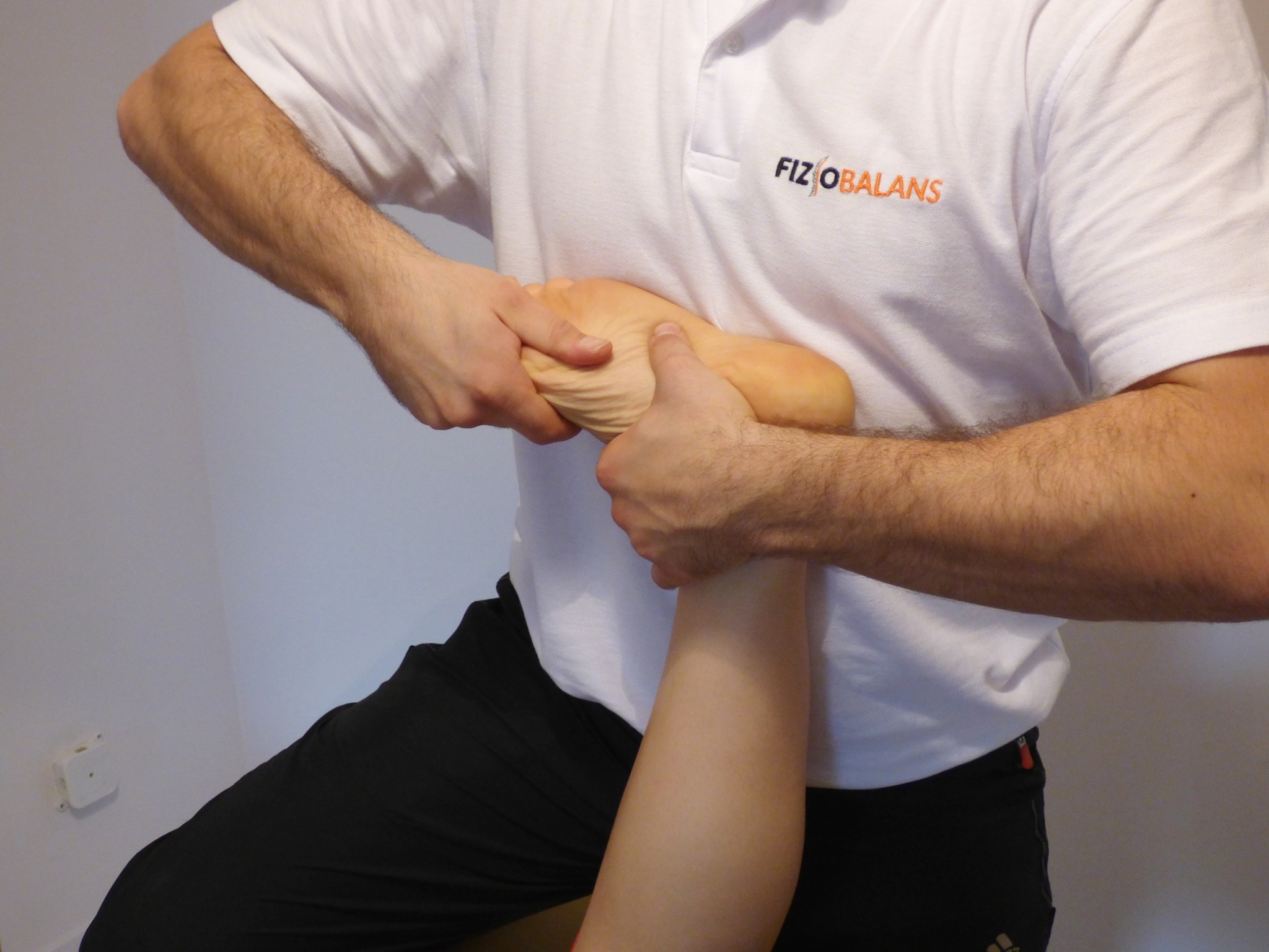 Masaż stopy wykonywany przez osobę w białej koszulce z logo 'FIZIOBALANS', widoczne czarne spodnie i fragment ściany.