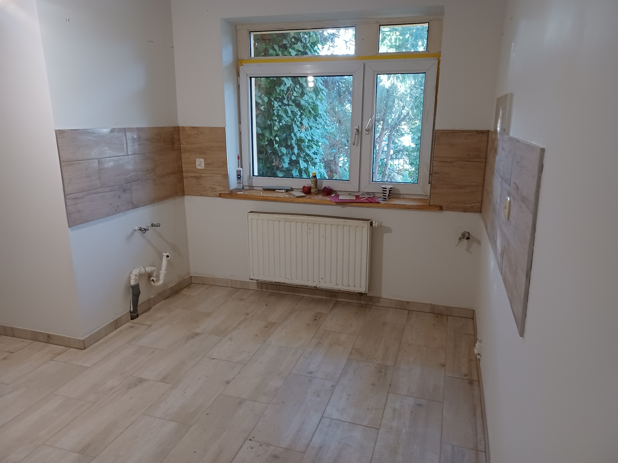 Wnętrze kuchni w trakcie remontu: częściowo położone płytki imitujące drewno na ścianach, widoczne wyprowadzenia hydrauliczne, grzejnik pod oknem, płytki podłogowe w odcieniach beżu.