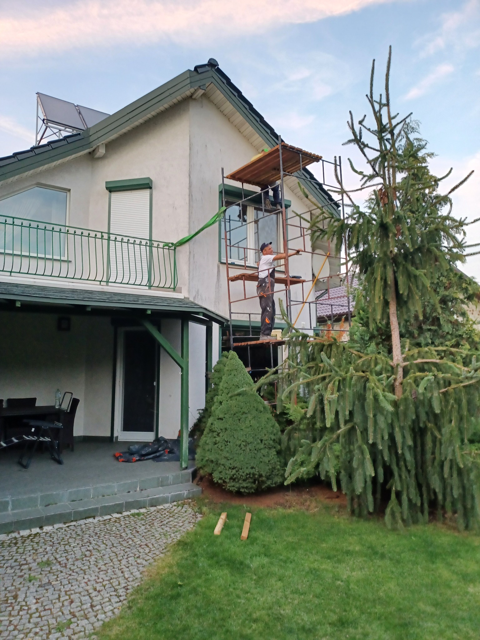 Pracownik na rusztowaniu podczas renowacji ściany zewnętrznej domu jednorodzinnego, widoczne panele solarne na dachu i iglaste drzewa w ogrodzie.