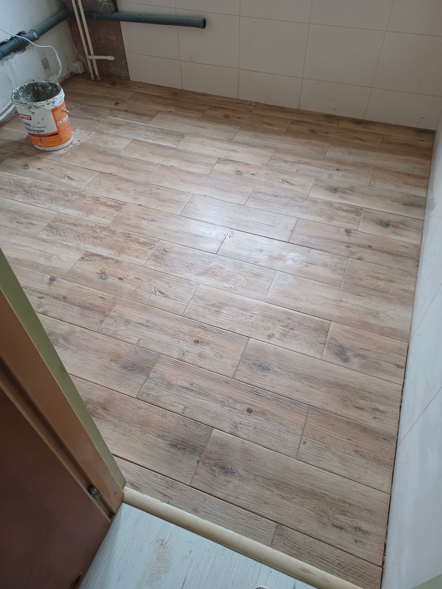 Ułożona podłoga z płytek imitujących drewno w jasnej łazience z białymi kafelkami na ścianach; widoczne rury i wiadro z farbą w rogu.