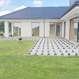 Przygotowanie podłoża pod taras: regulowane wsporniki ułożone na trawie, widoczny grill i taczka w tle, nowoczesny dom z dużymi oknami.