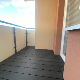 Nowoczesny balkon z ciemną, kompozytową podłogą, jasnymi ściankami bocznymi i czarną balustradą, widok z góry na fragment nieba.