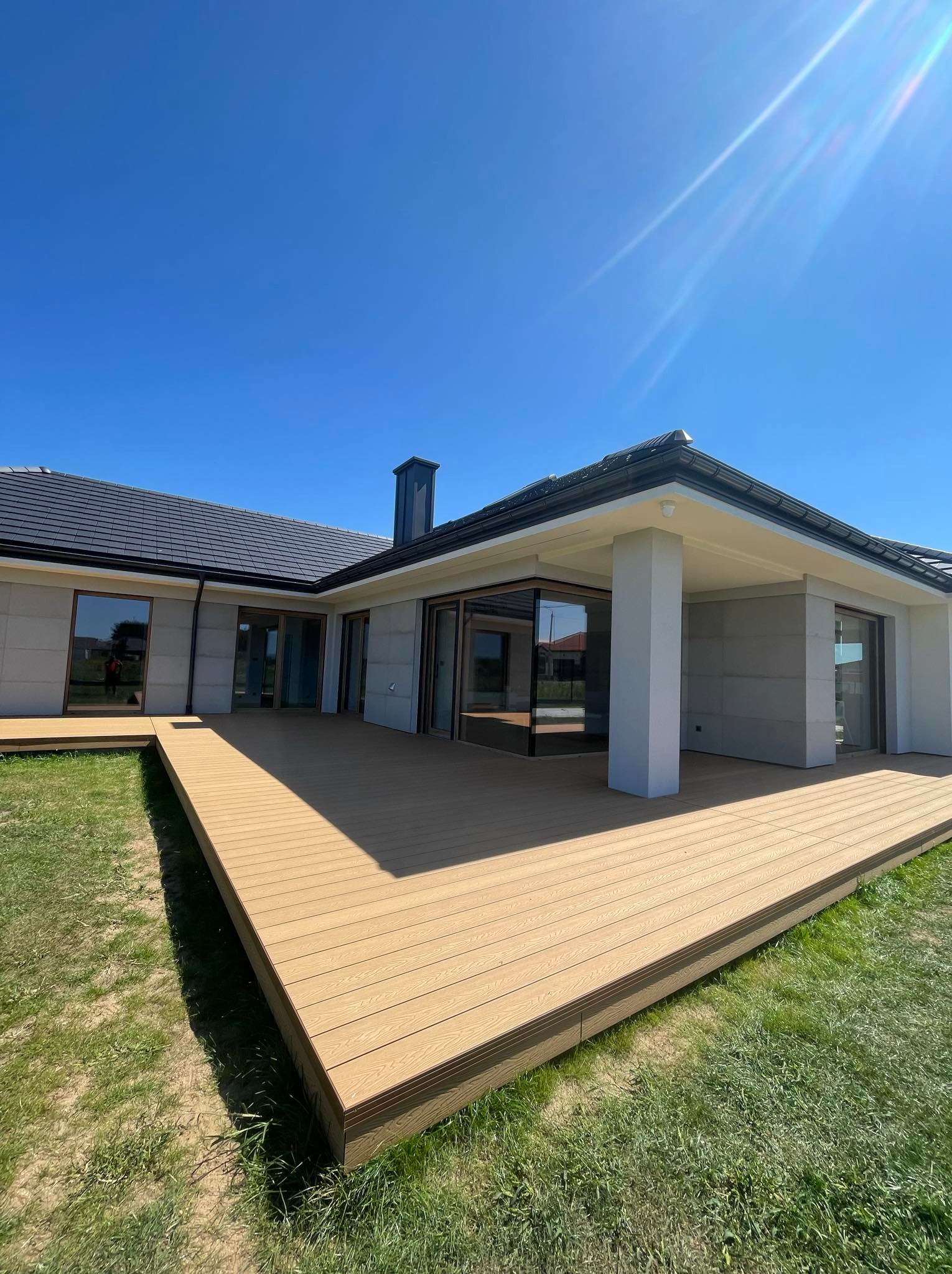 Nowoczesny dom z dużym tarasem z desek w kolorze jasnego drewna, widok z trawnika w słoneczny dzień.