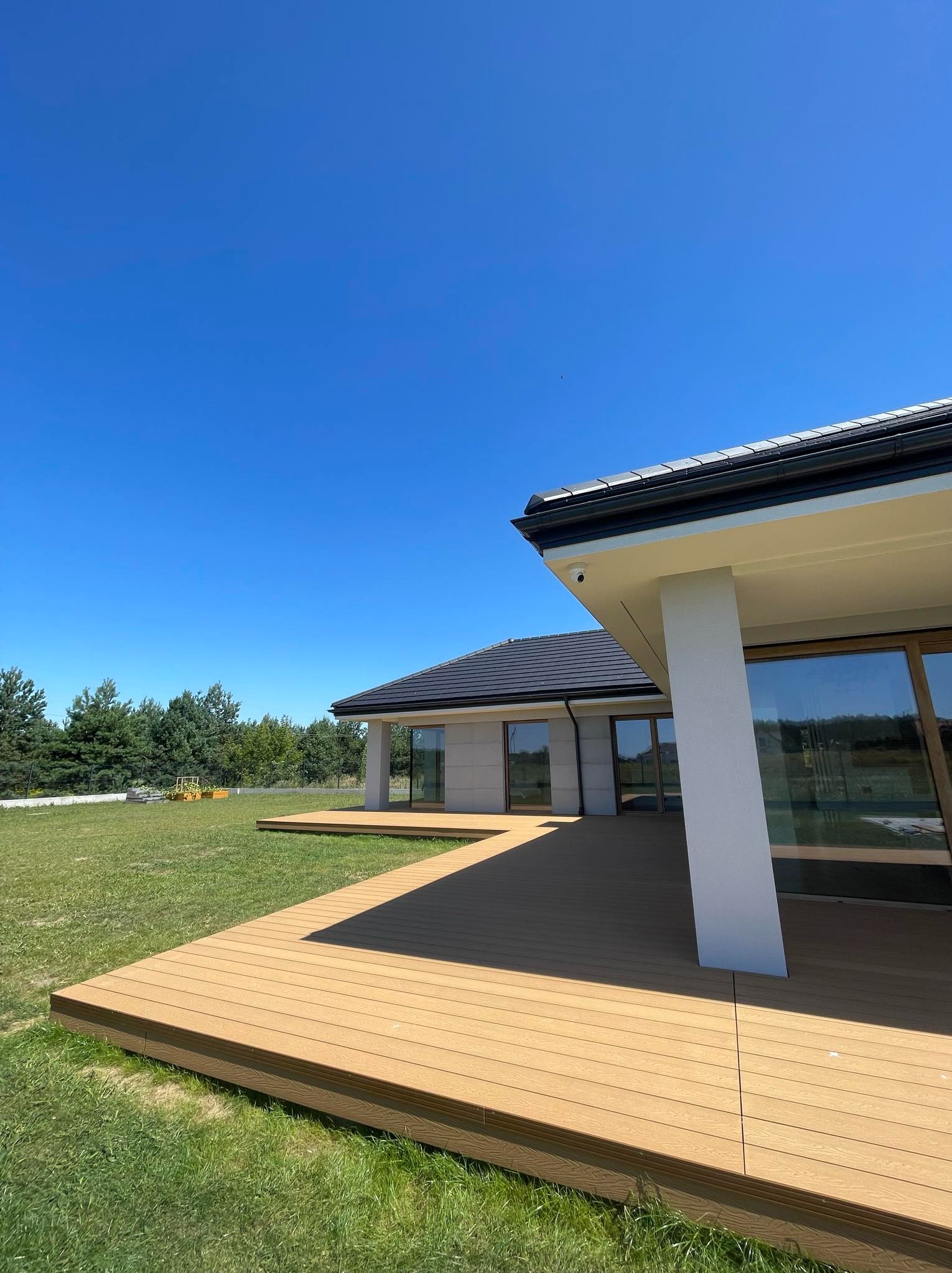Nowoczesny dom z rozległym tarasem z desek kompozytowych w kolorze jasnego drewna, widok z niskiej perspektywy na tle błękitnego nieba i zielonego trawnika.