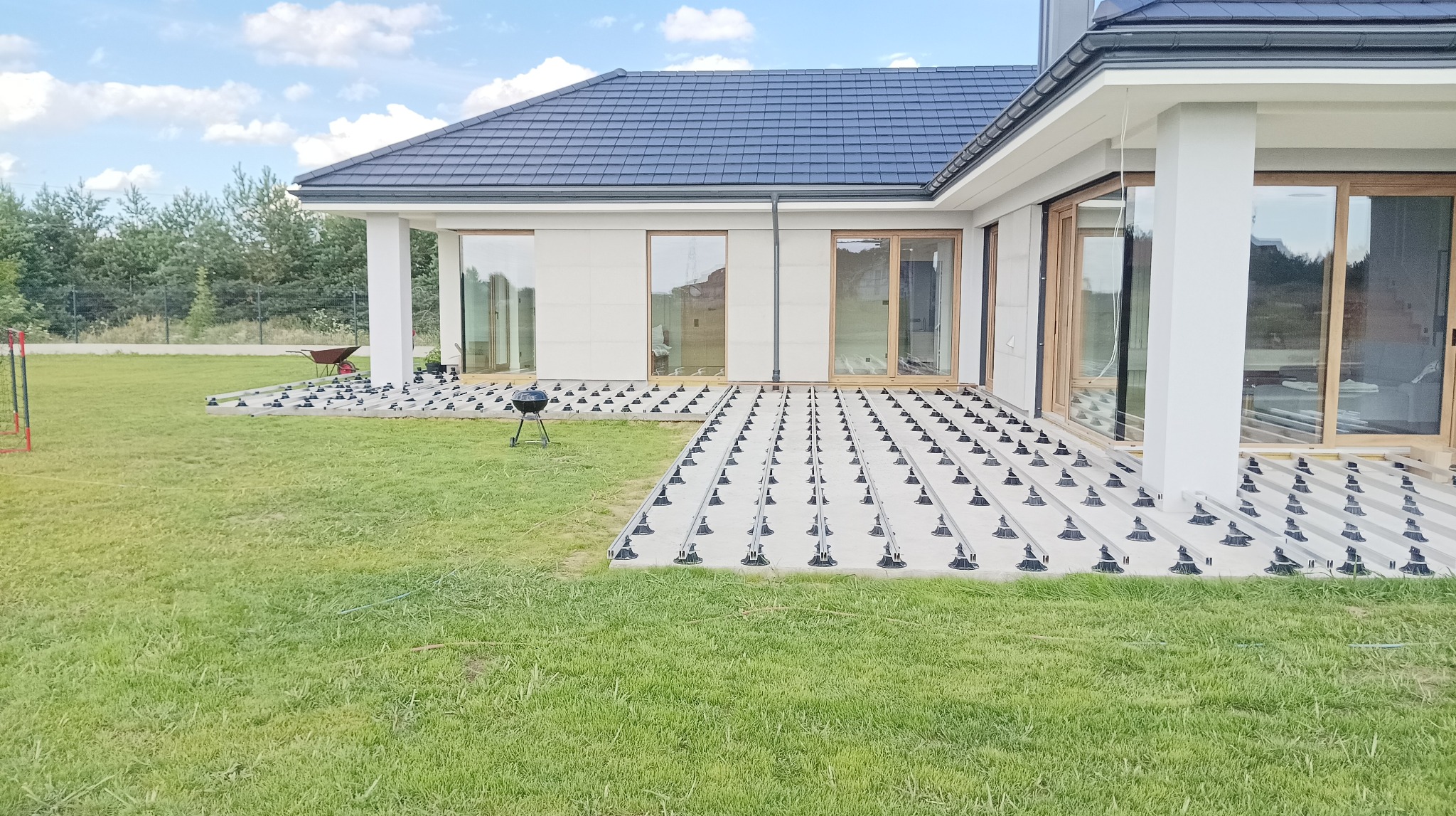 Przygotowanie podłoża pod taras: regulowane wsporniki ułożone na trawie, widoczny grill i taczka w tle, nowoczesny dom z dużymi oknami.