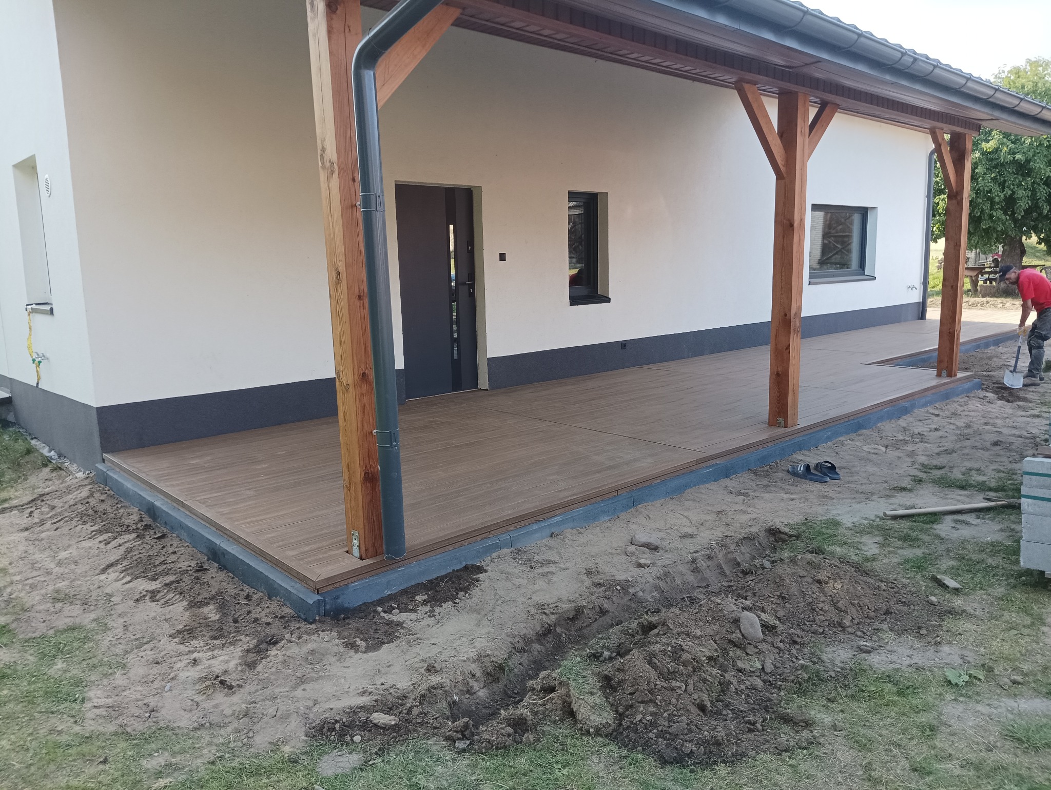 Jasny taras z desek kompozytowych przy białym domu z drewnianą konstrukcją zadaszenia, widoczne buty robocze oraz pracownik z łopatą w tle.