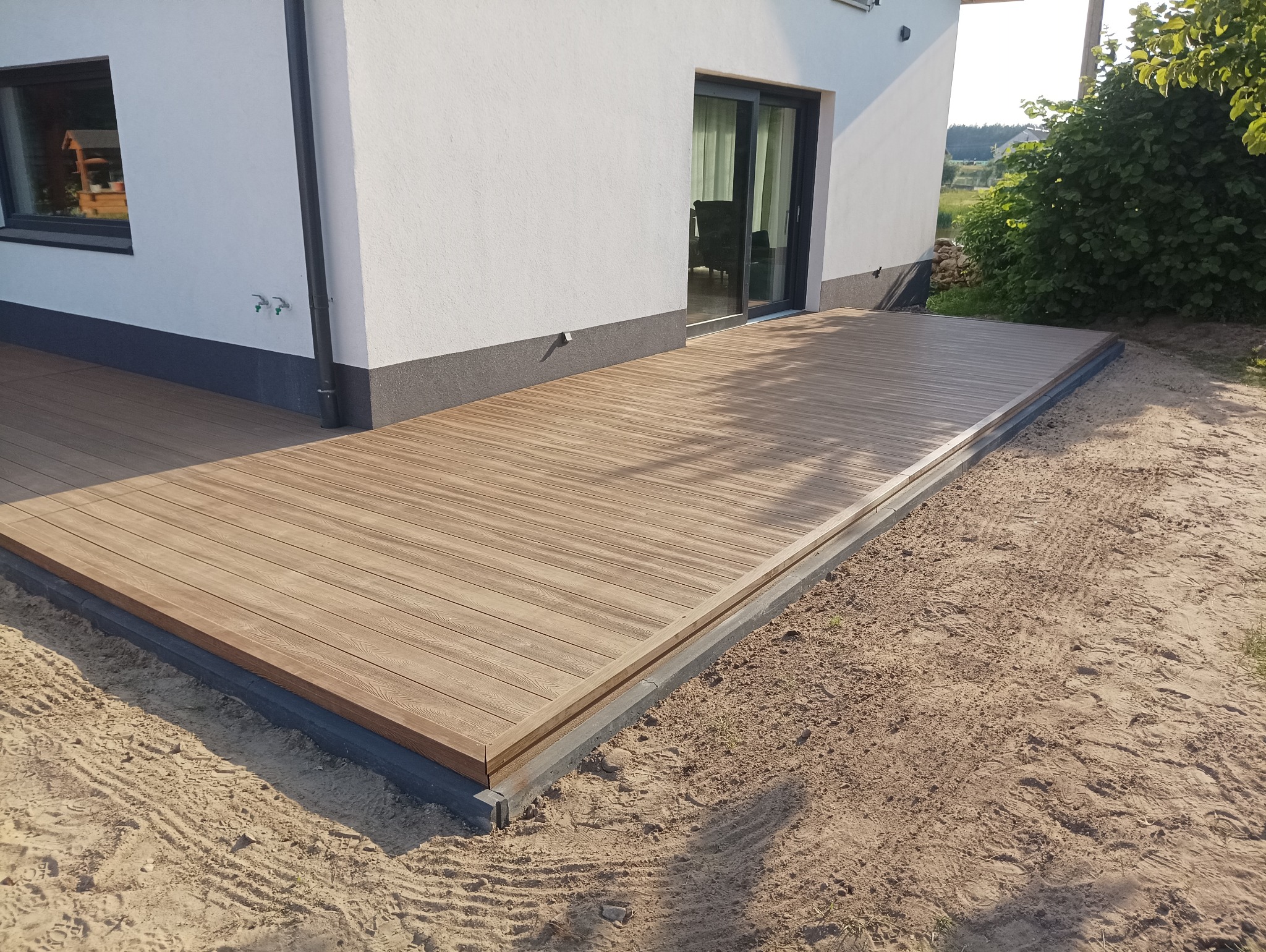 Nowoczesny taras z desek w kolorze jasnego drewna, obrzeże z szarej kostki, widok na dom z przesuwnymi drzwiami balkonowymi i ogród w tle.