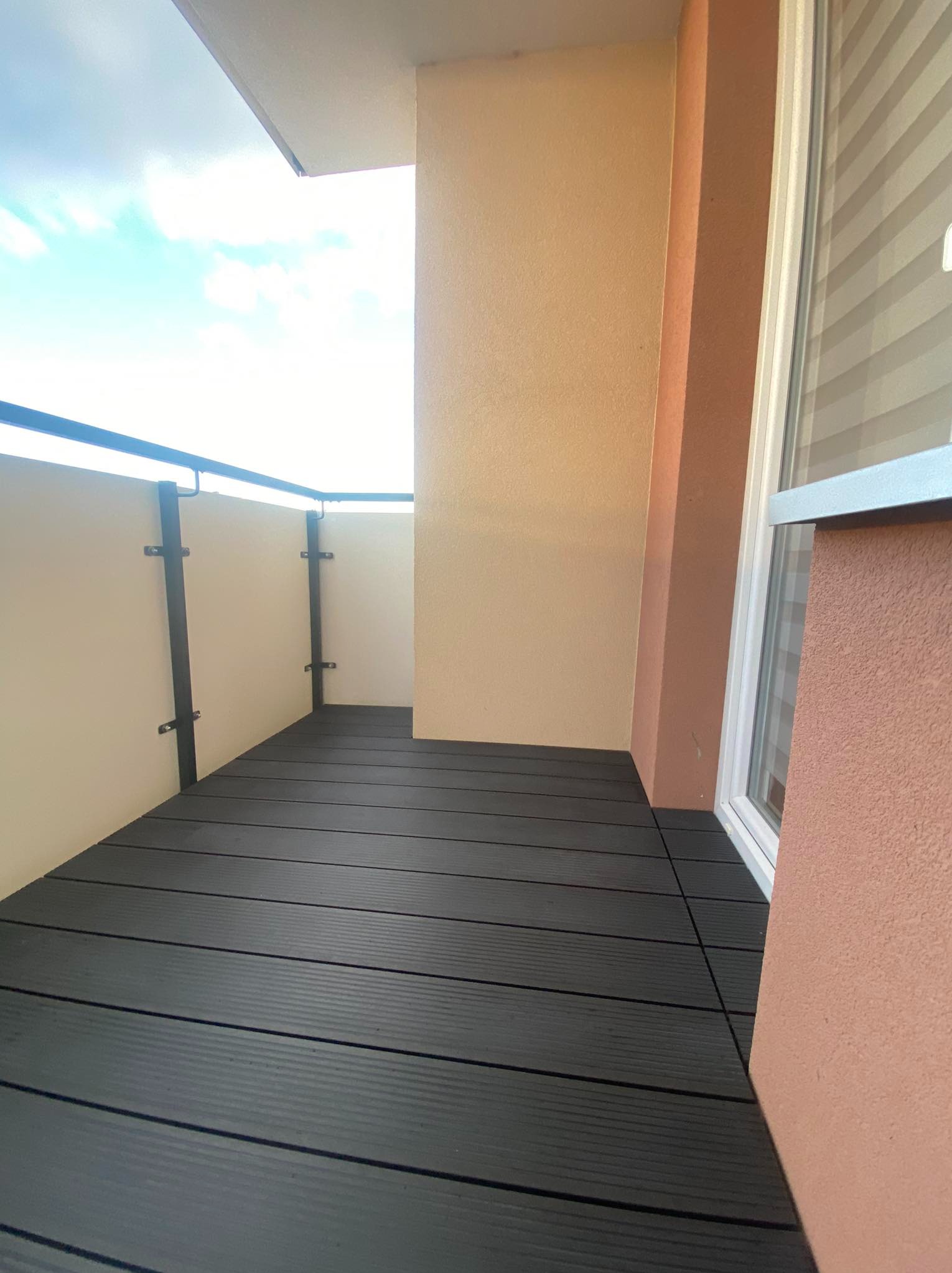 Nowoczesny balkon z ciemną, kompozytową podłogą, jasnymi ściankami bocznymi i czarną balustradą, widok z góry na fragment nieba.