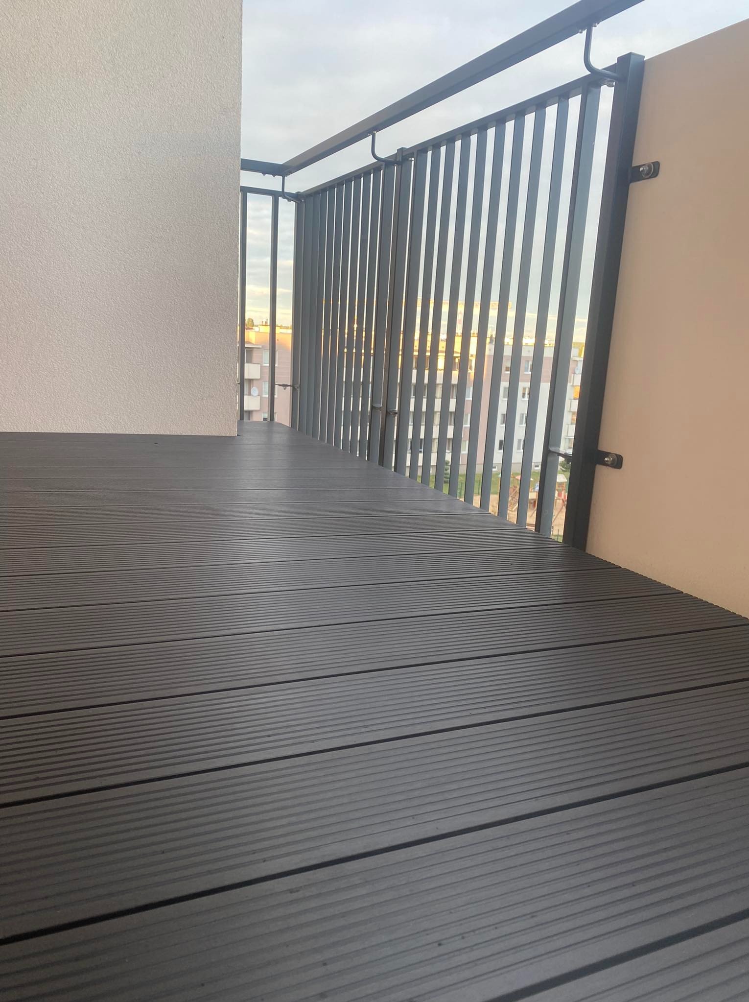 Nowoczesny balkon z szarą podłogą z desek kompozytowych i metalową balustradą z pionowych szczebli, widok na zabudowę miejską w tle.