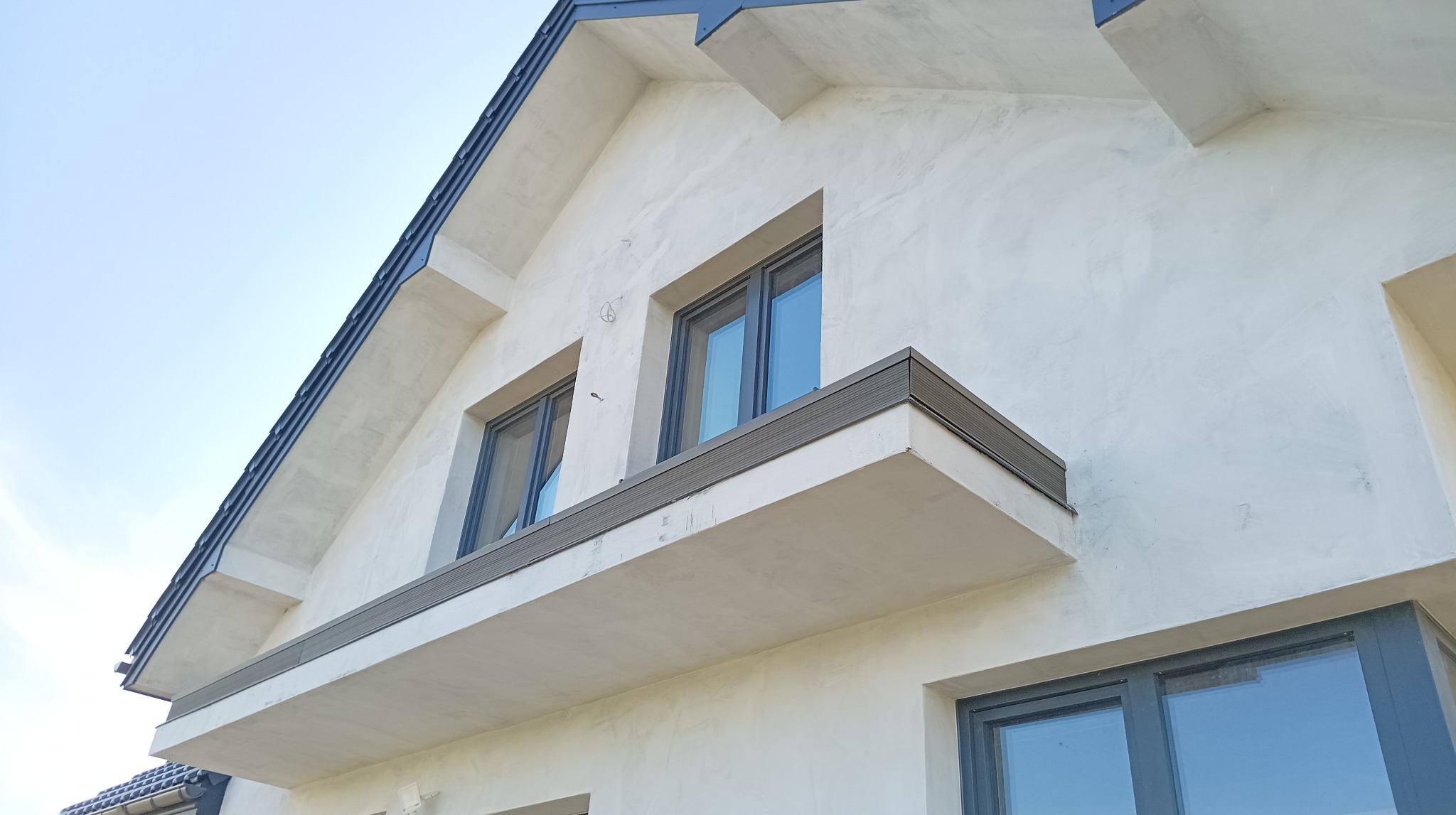 Nowoczesny balkon z szarą kompozytową deską elewacyjną, widok z dołu na jasną elewację budynku i niebieskie niebo.