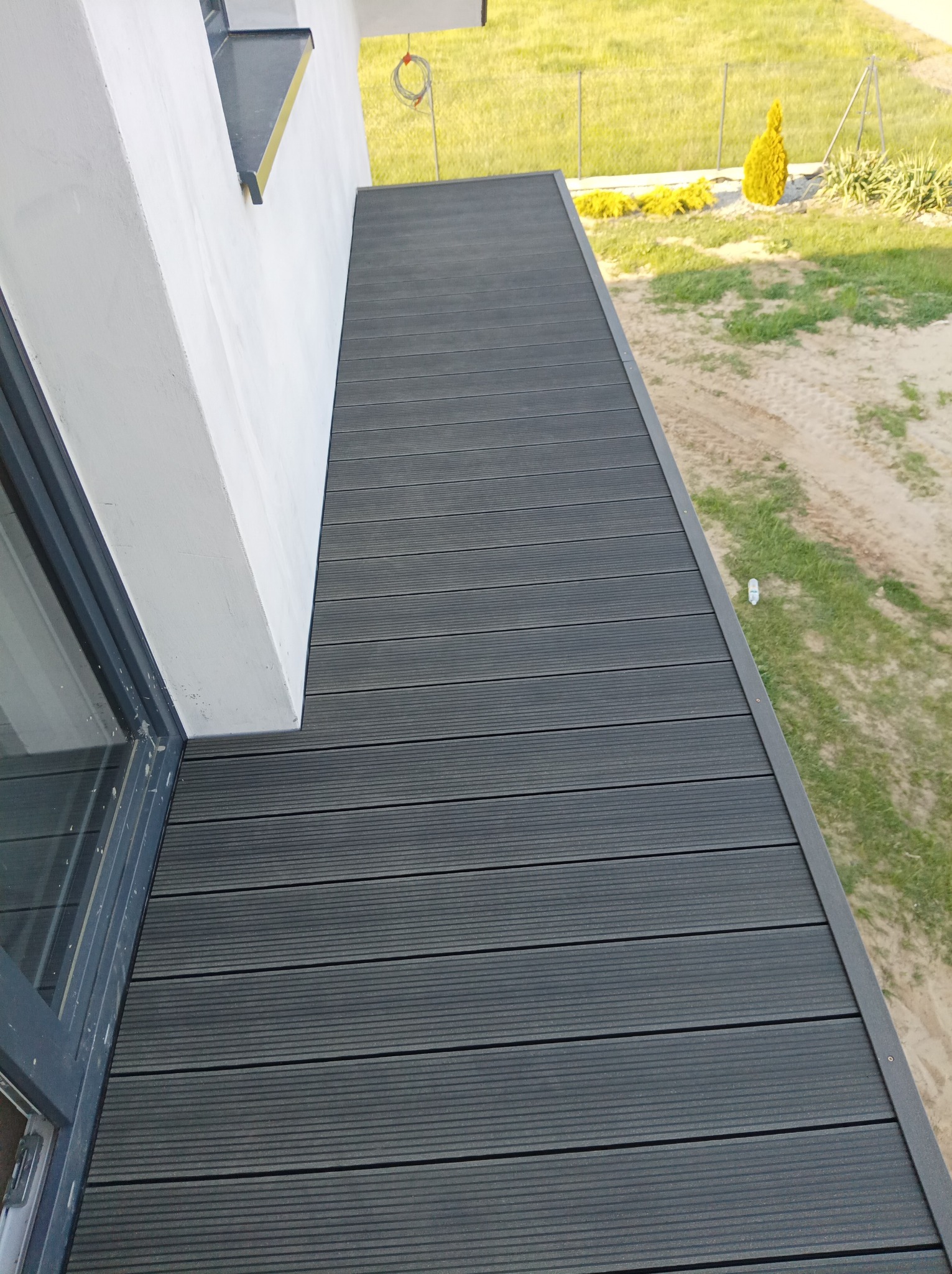 Nowoczesny balkon wykończony szarą deską kompozytową, widok z góry na taras z elementami zieleni w tle.