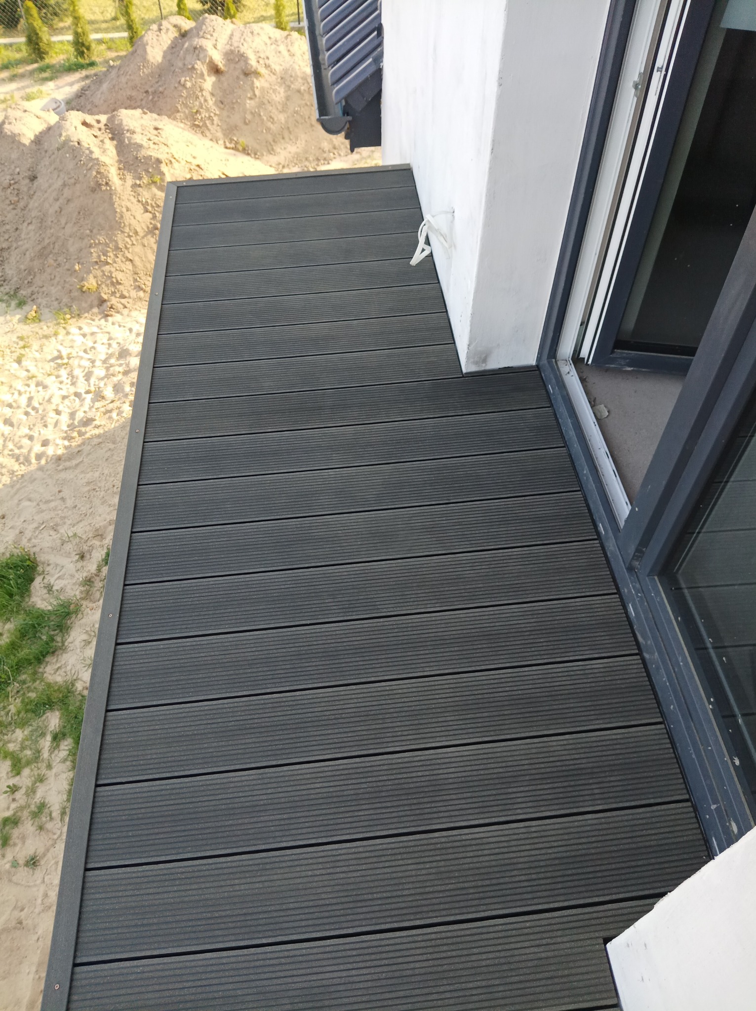 Nowoczesny taras z desek kompozytowych w kolorze grafitowym, widok z góry, z widocznym fragmentem okna i stertą piasku w tle.