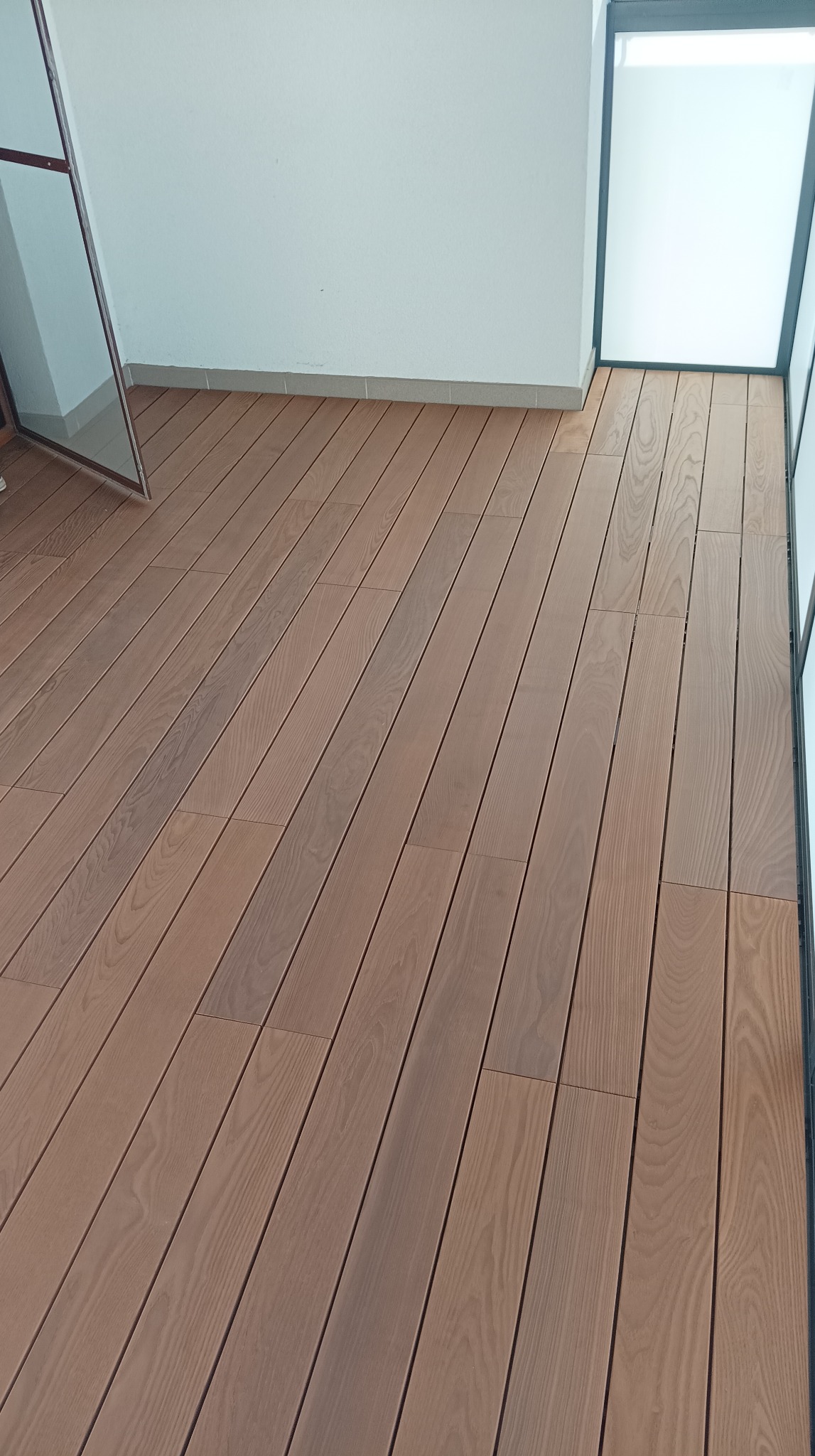 Jasnobrązowe deski tarasowe kompozytowe ułożone na balkonie, widoczne szklane drzwi balkonowe.