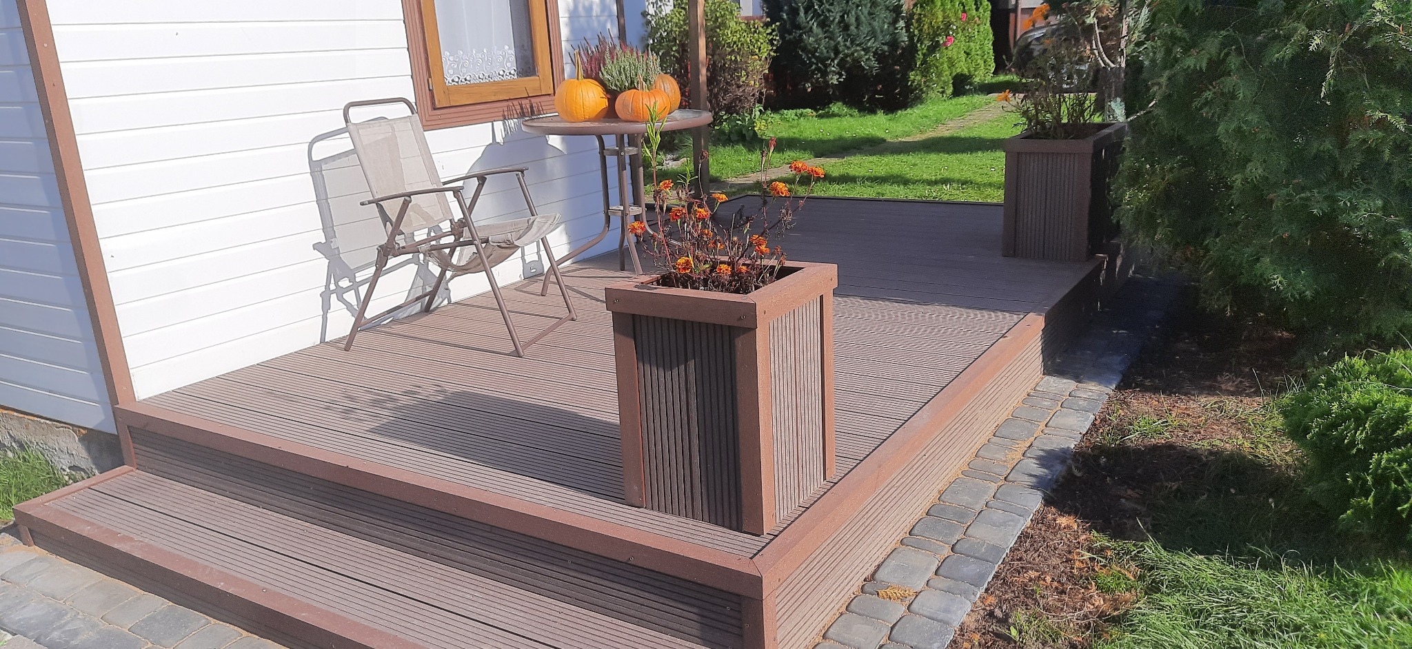 Brązowy taras z desek kompozytowych z dwoma schodkami, donicami z kwiatami i stolikiem z dyniami, obok biały domek z brązowymi ramami okiennymi.