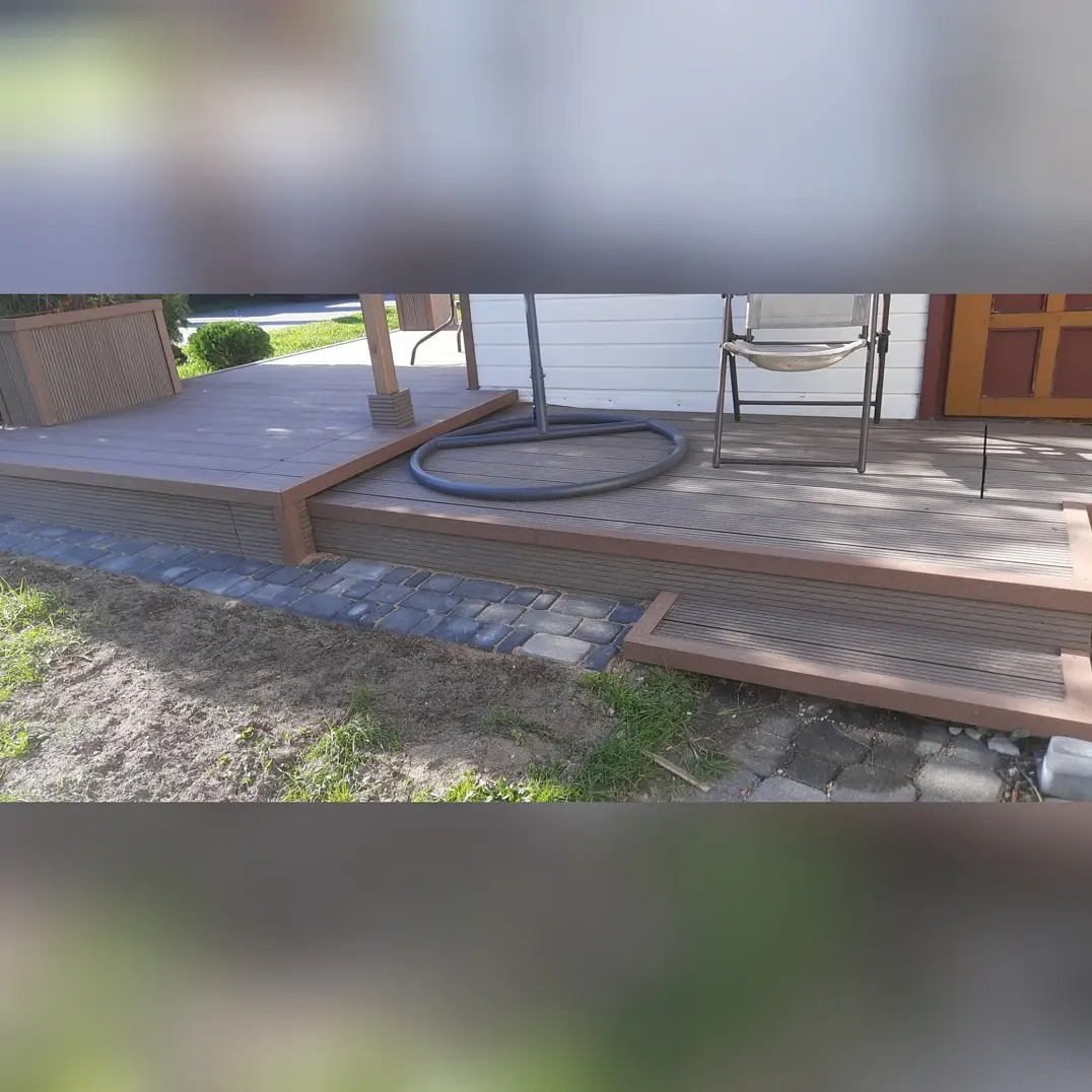 Wykończony taras z desek w odcieniach brązu, obok ułożona kostka brukowa, widoczne krzesło ogrodowe i element huśtawki.