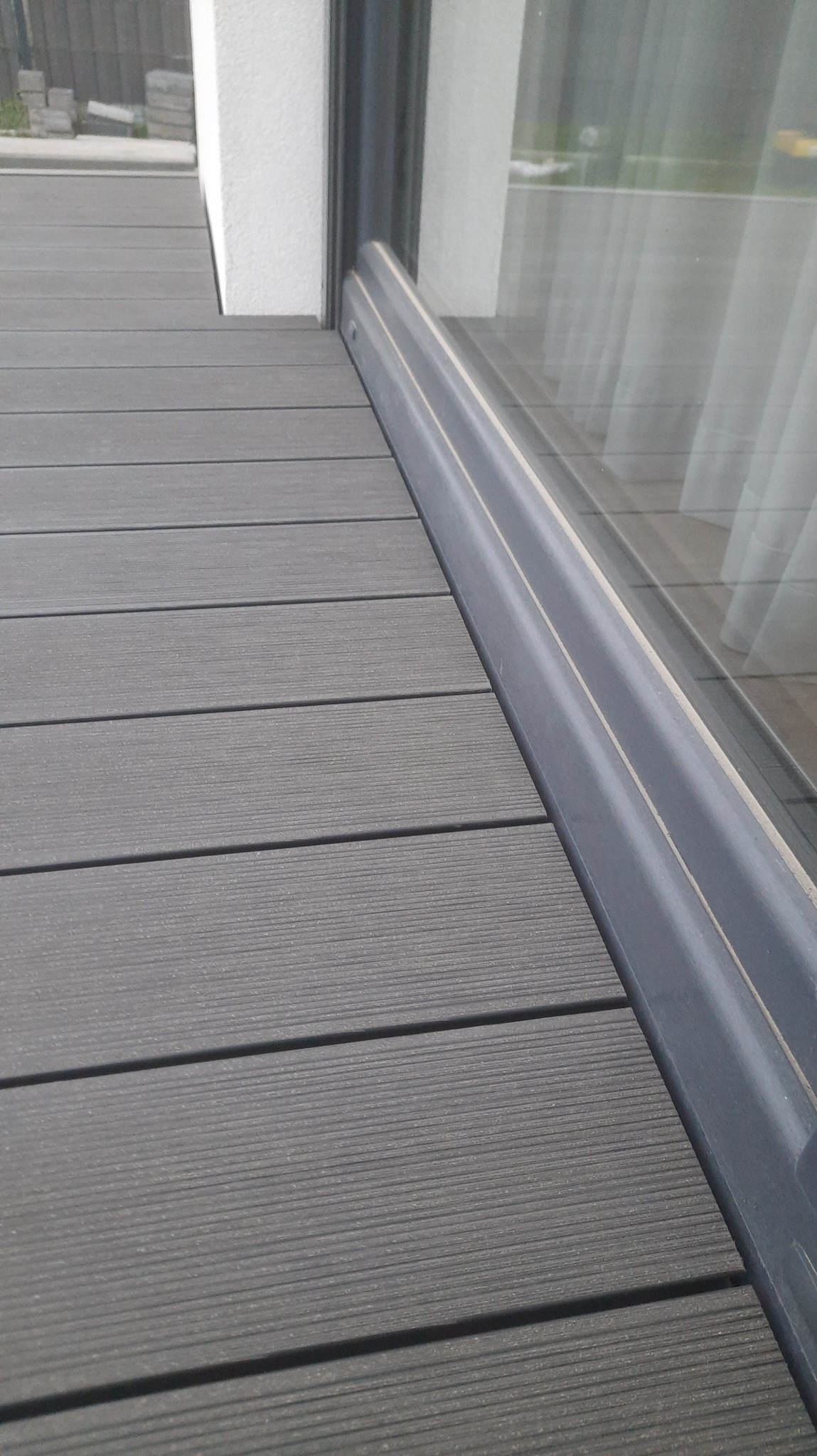 Szare deski tarasowe kompozytowe ułożone przy oknie balkonowym, widoczna faktura desek i profil okna.