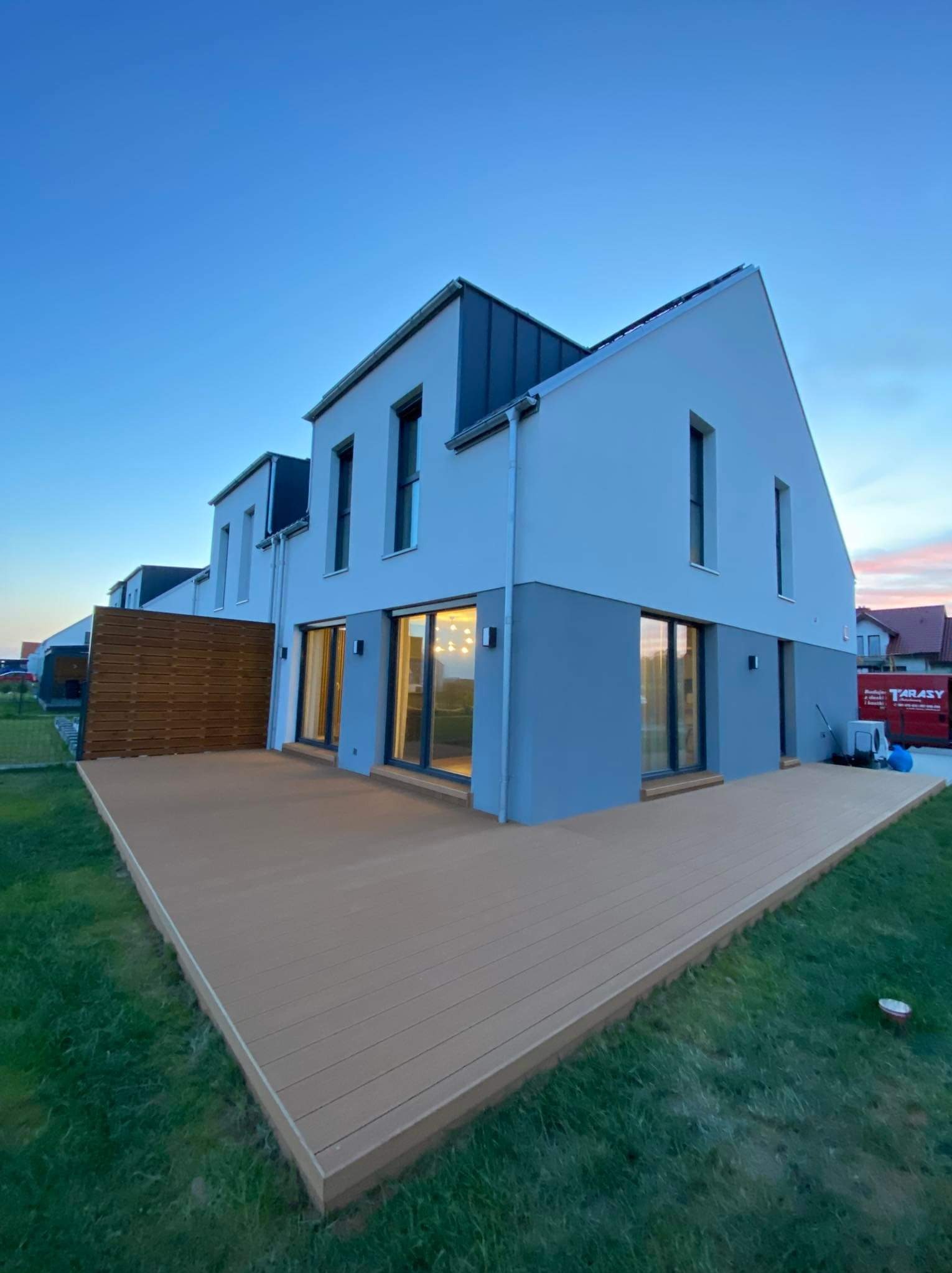 Nowoczesny dom z szarą elewacją i brązowym tarasem z desek kompozytowych, widok z trawnika o zmierzchu.