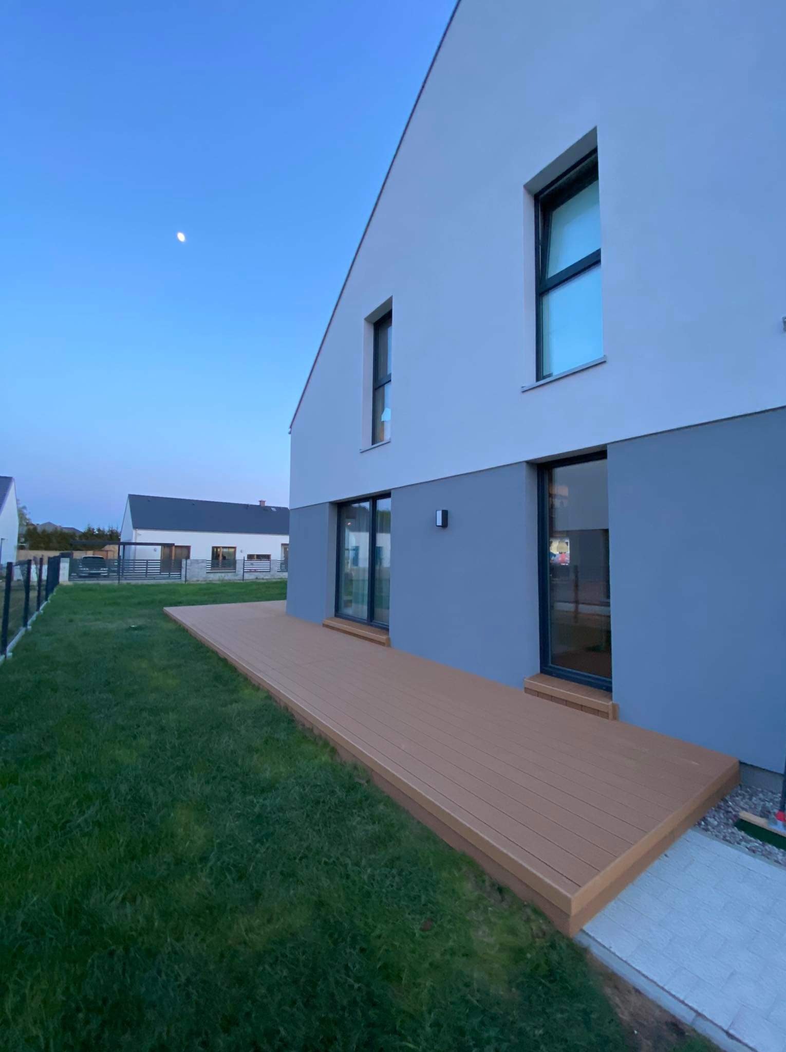 Nowoczesny dom z brązowym tarasem z desek kompozytowych, widok na trawnik i błękitne niebo o zmierzchu.