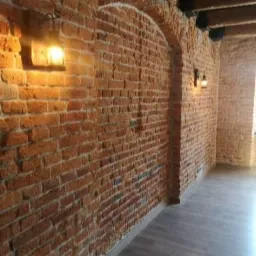 Wnętrze z odsłoniętą cegłą, oświetlone kinkietami z ciepłym światłem, ukazujące łukowate przejście i drewniany sufit.