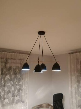 Czarna lampa wisząca z czterema kloszami w kształcie kopuł, zamontowana na drewnianej listwie, oświetlająca wnętrze z firankami i biurowym krzesłem.