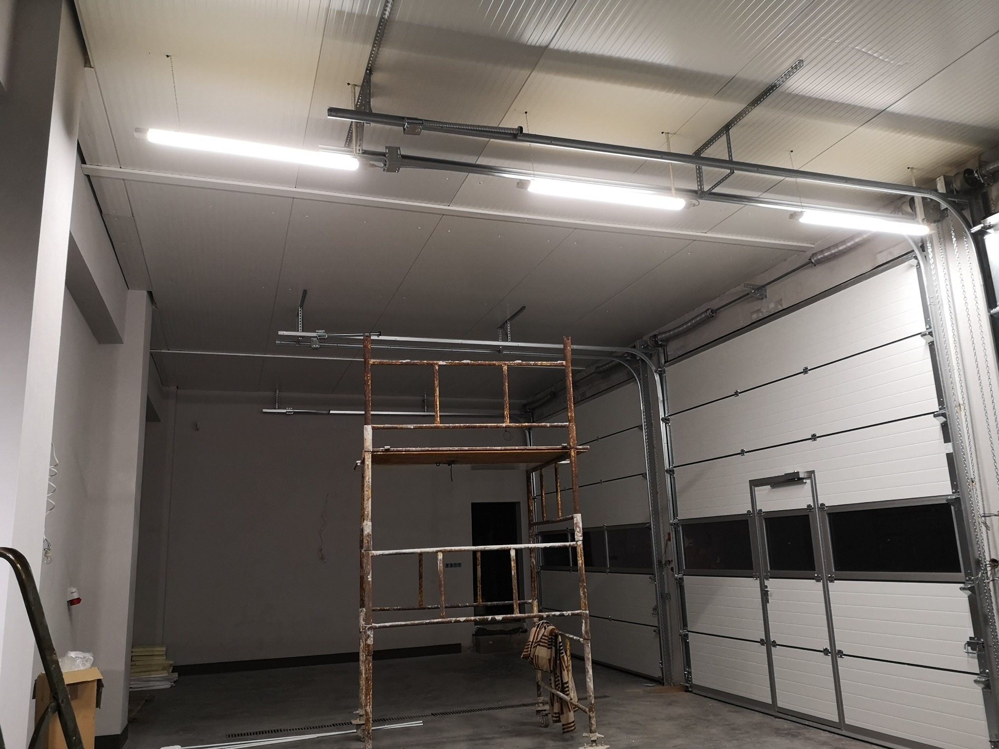 Wnętrze hali z zamontowanym oświetleniem LED, widoczne rusztowanie, segmentowa brama garażowa i surowe wykończenie ścian.