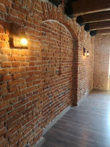 Wnętrze z odsłoniętą cegłą, oświetlone kinkietami z ciepłym światłem, ukazujące łukowate przejście i drewniany sufit.