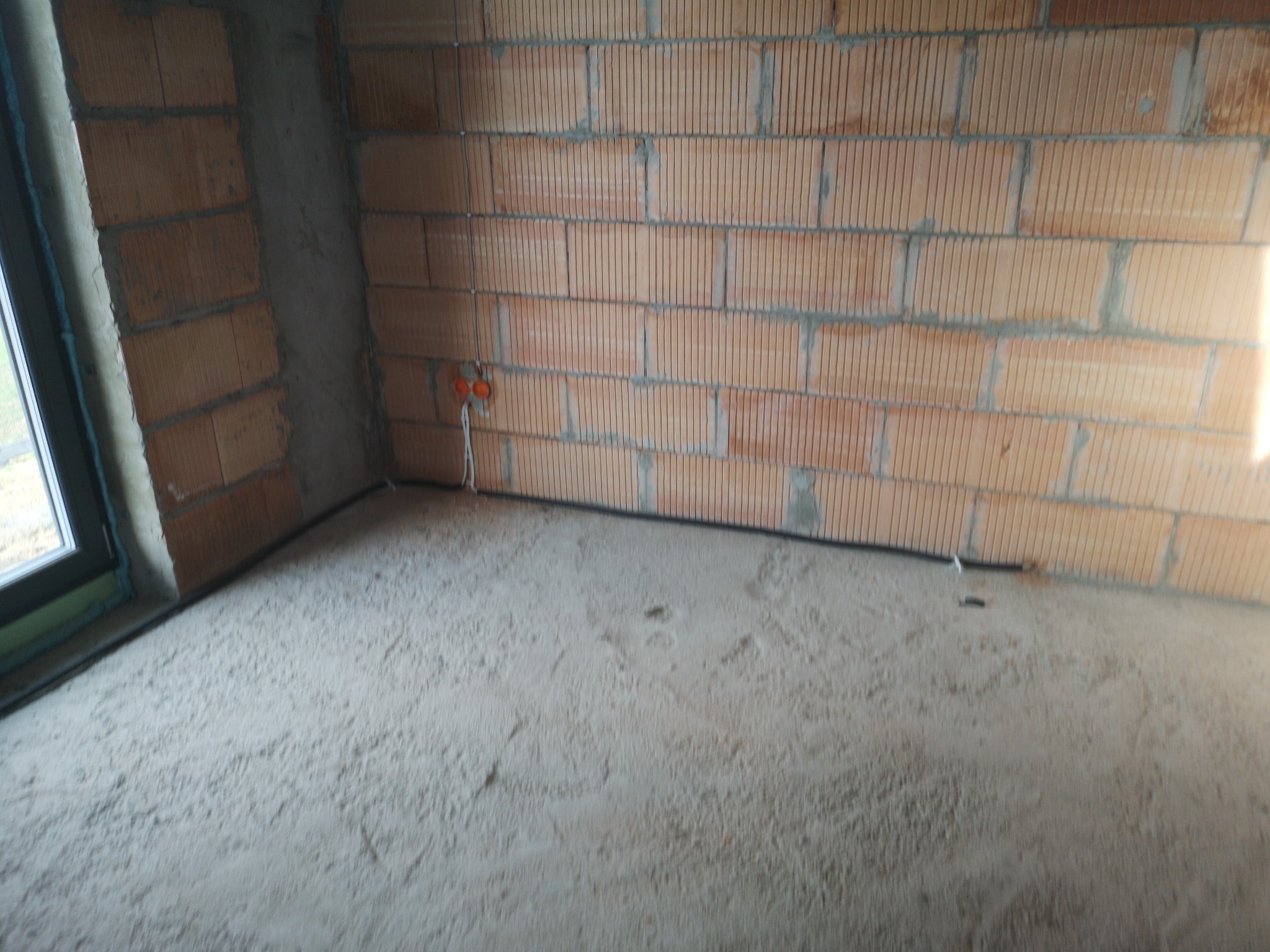 Surowy stan wnętrza z ceglanymi ścianami, widoczne puszki elektryczne i przewody wystające ze ściany, okno z lewej strony.