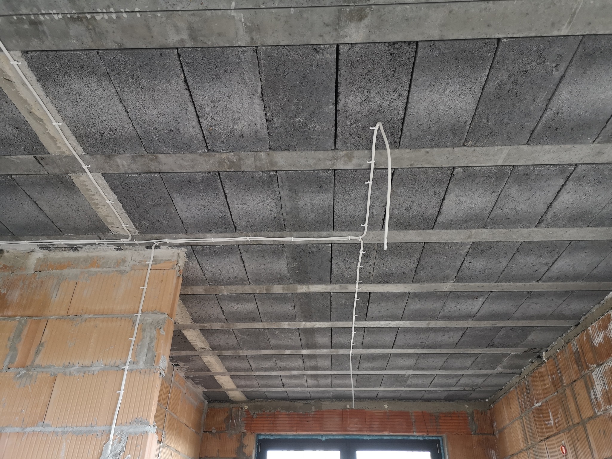 Surowy strop z betonowych bloczków z rozprowadzonymi przewodami elektrycznymi przymocowanymi klamrami, widok na budowę w stanie surowym.