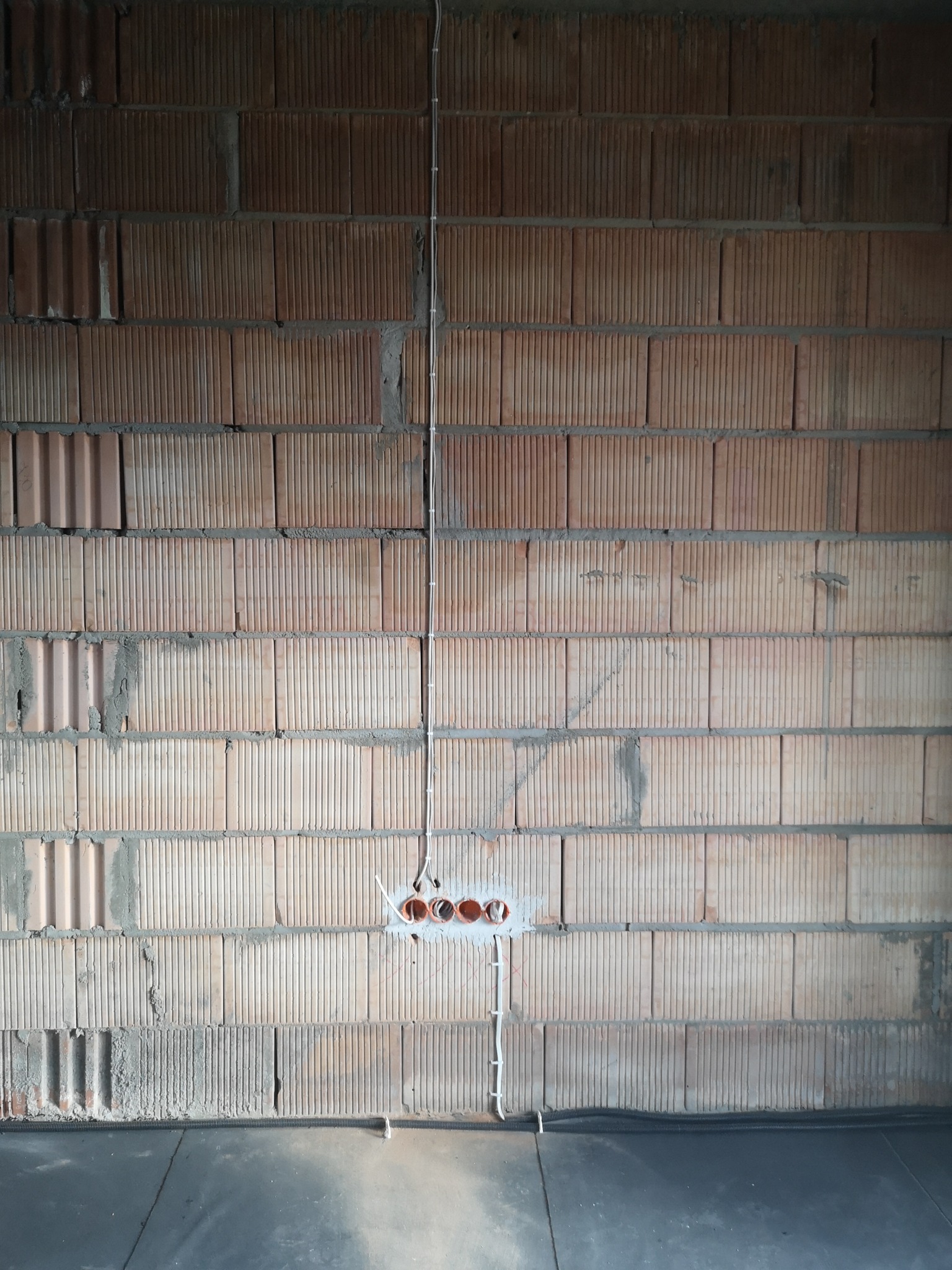 Surowa ściana z cegły dziurawki z widocznymi przewodami elektrycznymi w peszlu i puszką pod gniazdka, instalacja w trakcie montażu.