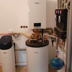 Kotłownia z białym piecem gazowym Vaillant, zasobnikiem ciepłej wody i zmiękczaczem wody Blues, widoczne rury i instalacje.