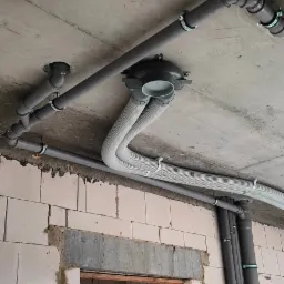 Szare rury wentylacyjne i hydrauliczne zamontowane pod surowym betonowym stropem, widoczne fragmenty ścian z białego bloczka w surowym stanie.