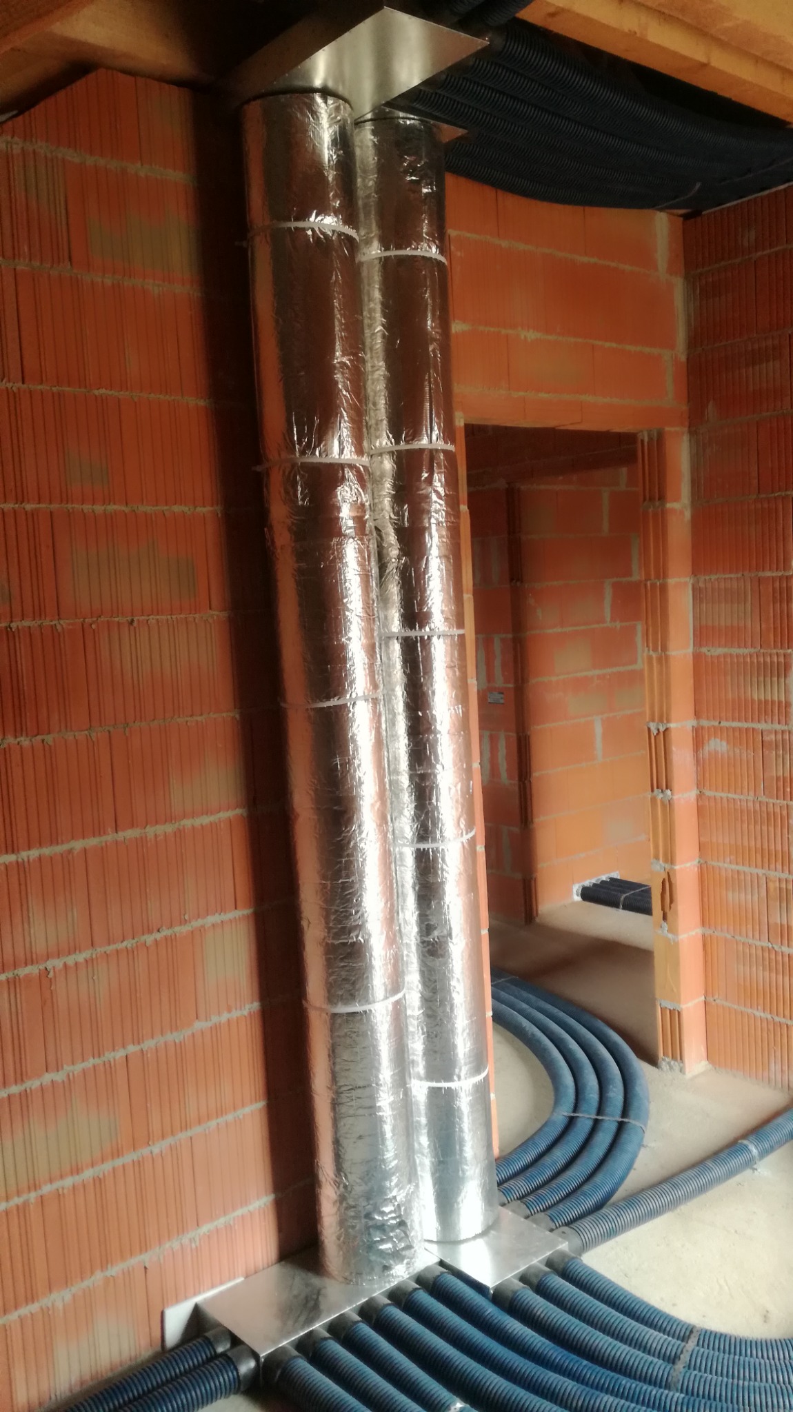 Dwa pionowe, izolowane przewody wentylacyjne z folii aluminiowej, zamontowane w surowym wnętrzu z ceglanymi ścianami, z widocznymi niebieskimi rurami wentylacyjnymi ułożonymi na podłodze.