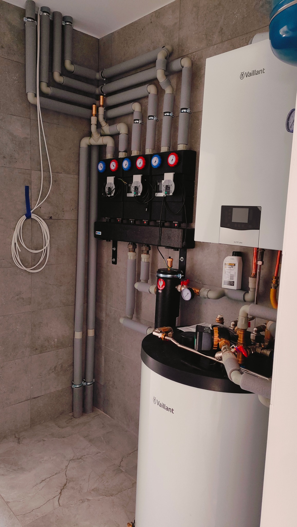 Węzeł cieplny z kotłem gazowym Vaillant, rozdzielaczami z manometrami i orurowaniem w pomieszczeniu z szarą płytką imitującą marmur.