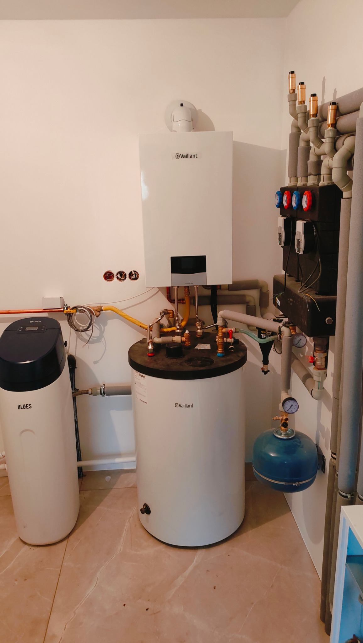 Kotłownia z białym piecem gazowym Vaillant, zasobnikiem ciepłej wody i zmiękczaczem wody Blues, widoczne rury i instalacje.