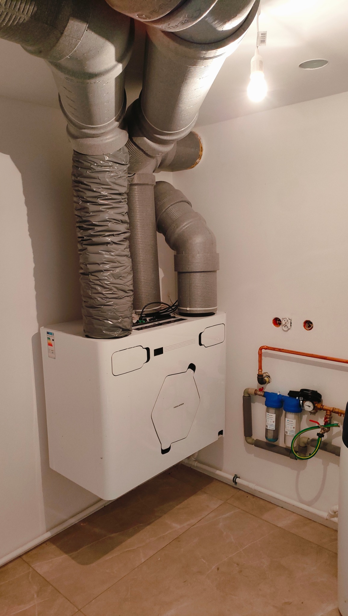 Biała jednostka centralna z szarymi przewodami wentylacyjnymi w pomieszczeniu z terakotą, widoczne rury miedziane i filtry wody.