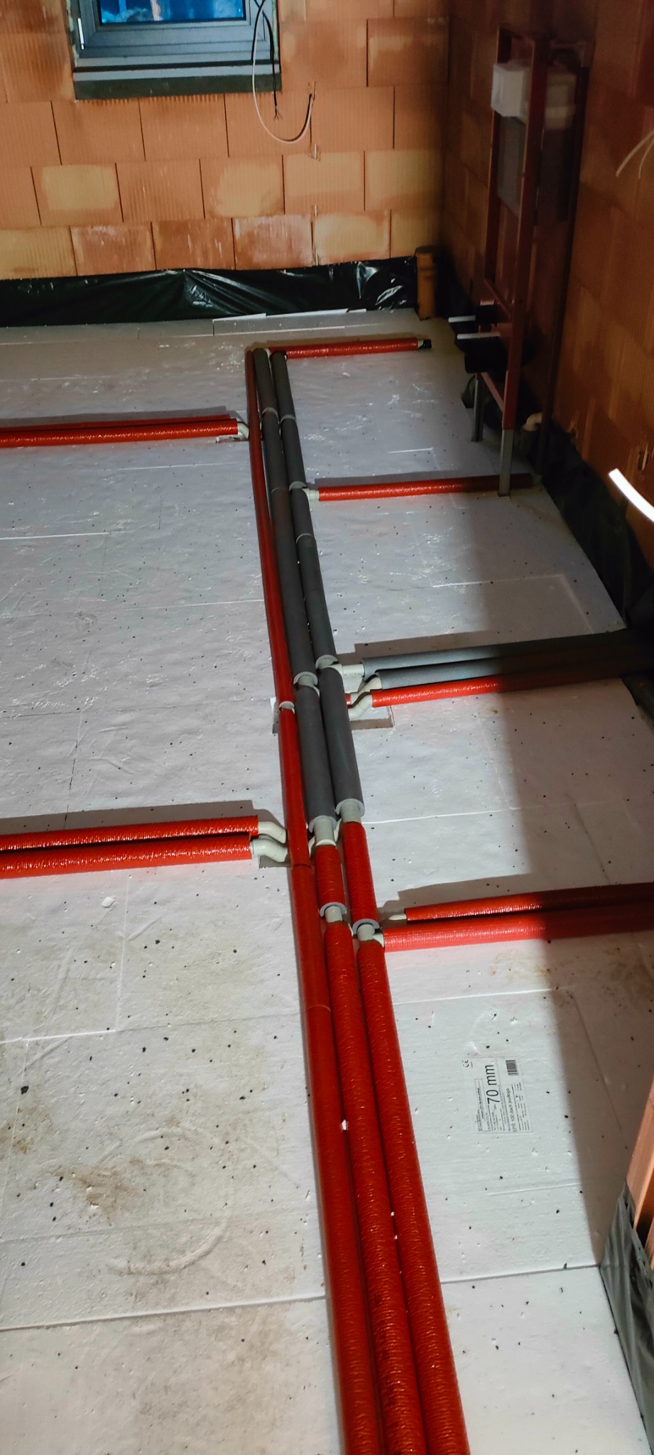 Czerwone rury ogrzewania podłogowego ułożone na białym styropianie, częściowo połączone z szarymi rurami w surowym stanie, widoczne fragmenty ściany z cegły dziurawki i okno.