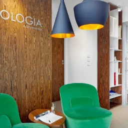 Wnętrze gabinetu z drewnianą ścianą z napisem 'Podologia A.Szymańska', zielonymi fotelami, stolikiem z gazetą i wodą, wiszącymi lampami z złotym wnętrzem i regałem z książkami w tle