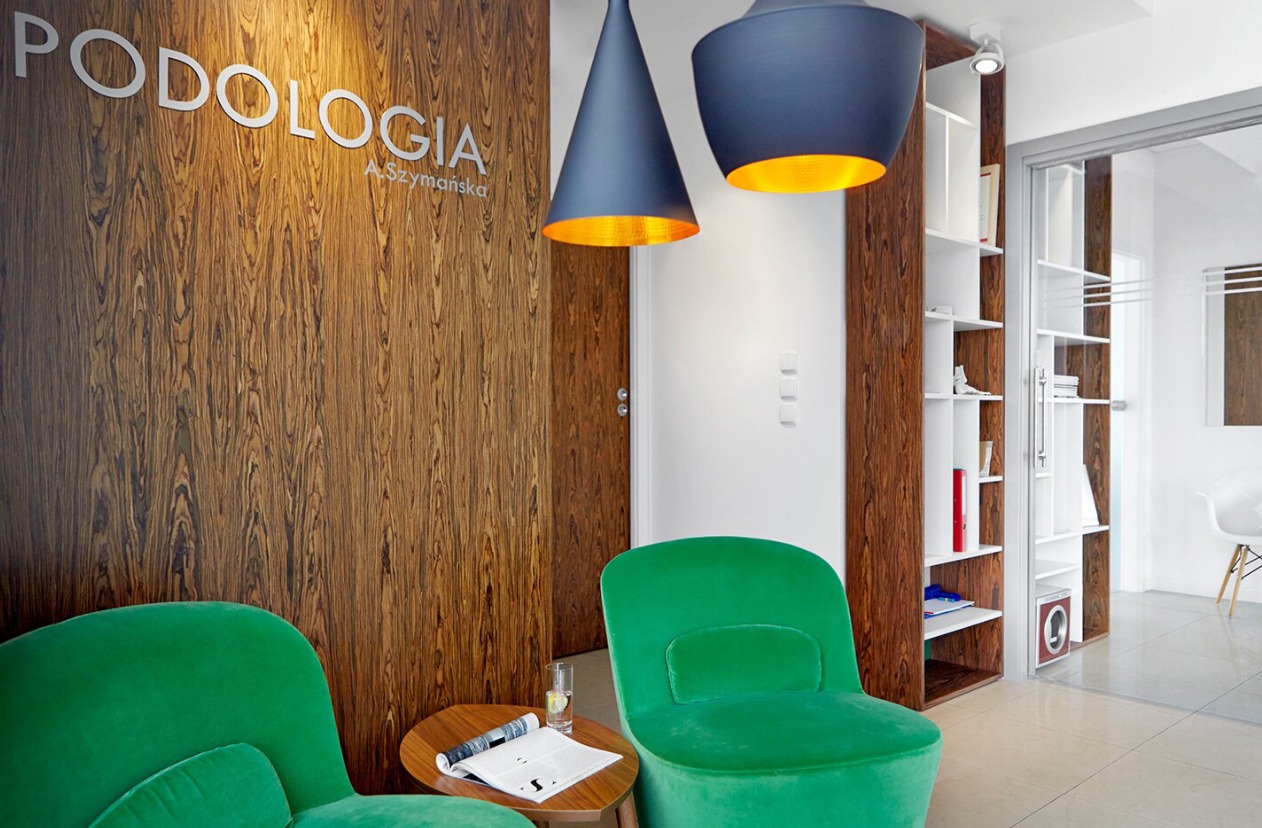 Wnętrze gabinetu z drewnianą ścianą z napisem 'Podologia A.Szymańska', zielonymi fotelami, stolikiem z gazetą i wodą, wiszącymi lampami z złotym wnętrzem i regałem z książkami w tle
