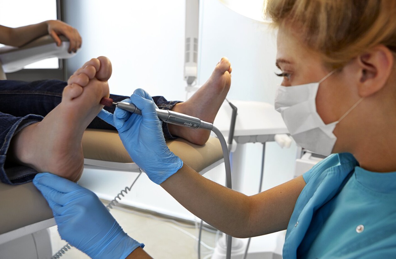 Pedicure frezarkowy w trakcie wykonywania: specjalistka w masce i rękawiczkach opracowuje skórę stopy klienta frezem.