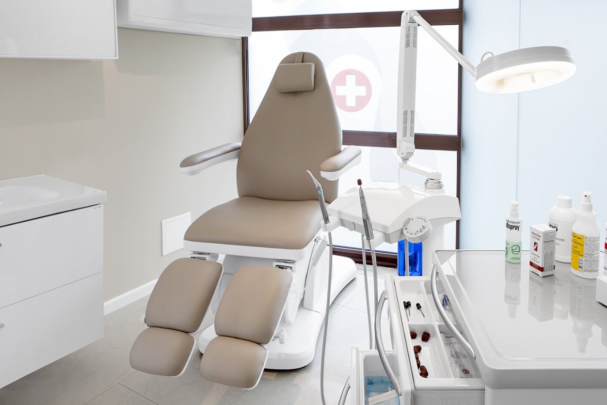 Nowoczesny gabinet zabiegowy z beżowym fotelem, lampą operacyjną i sterylnymi narzędziami, widok na okno z symbolem plusa.