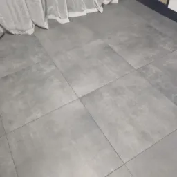 Szare, matowe płytki podłogowe ułożone w kwadraty, widoczne fragmenty białych zasłon i półki z kosmetykami.