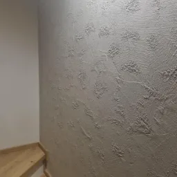 Ściana pokryta dekoracyjną, strukturalną gładzią w odcieniach szarości, przylegająca do drewnianych schodów.