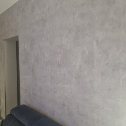 Krzysztof Tkacz KRISBUD Usługi remontowo-budowlane - Ściana wykończona szarą, betonową strukturą, widoczny fragment ościeżnicy drzwi i fragment granatowej sofy.