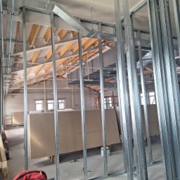 Krzysztof Tkacz KRISBUD Usługi remontowo-budowlane - Montaż metalowej konstrukcji szkieletowej ścian działowych w dużym pomieszczeniu z widoczną konstrukcją dachu i płytami gipsowo-kartonowymi, czerwony plecak na pierwszym planie.